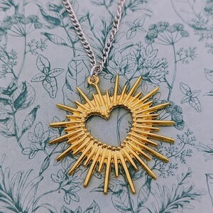 May include: A gold necklace with a large, sunburst-shaped pendant featuring a heart cutout. The pendant has a textured, beaded edge.