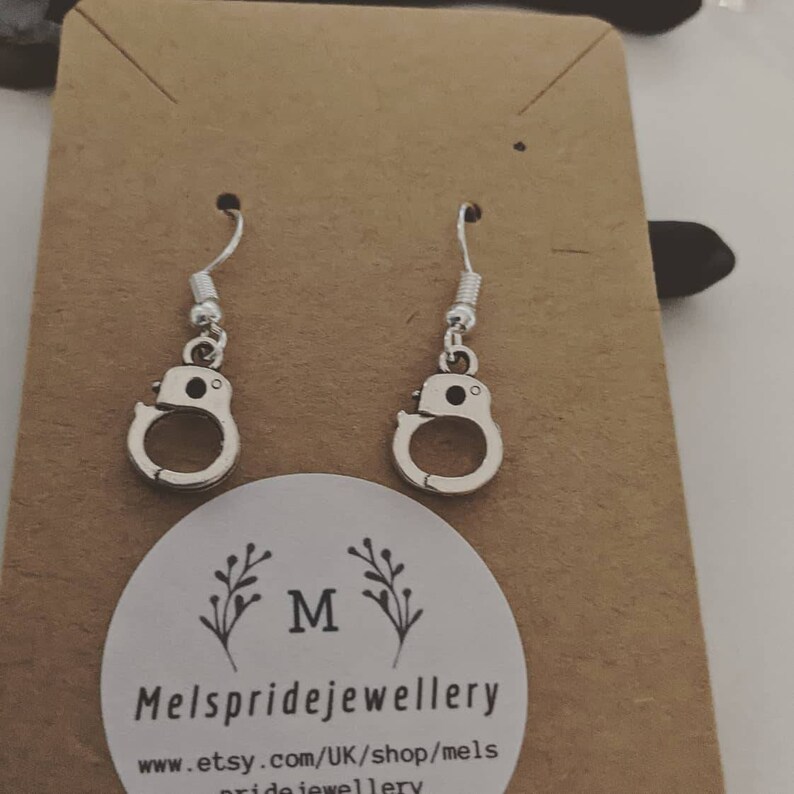 May include: Silver handcuff dangle earrings on a brown card with the text "Mels pride jewellery" and the website address "www.etsy.com/UK/shop/mels pride jewellery".