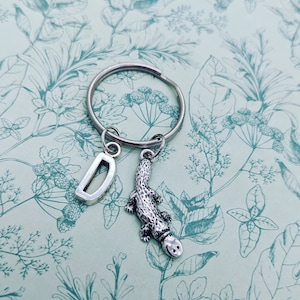 Op de afbeelding: Een zilveren sleutelhanger met een letter D-bedel en een vogelbekdier-bedel. De bedels zijn bevestigd aan een zilveren sleutelring. De achtergrond is een lichtblauw en groen bloemenpatroon.