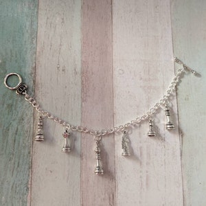 May include: A silver charm bracelet with a variety of chess piece charms, including a king, queen, knight, and pawn. The bracelet has a silver clasp and a silver toggle closure.