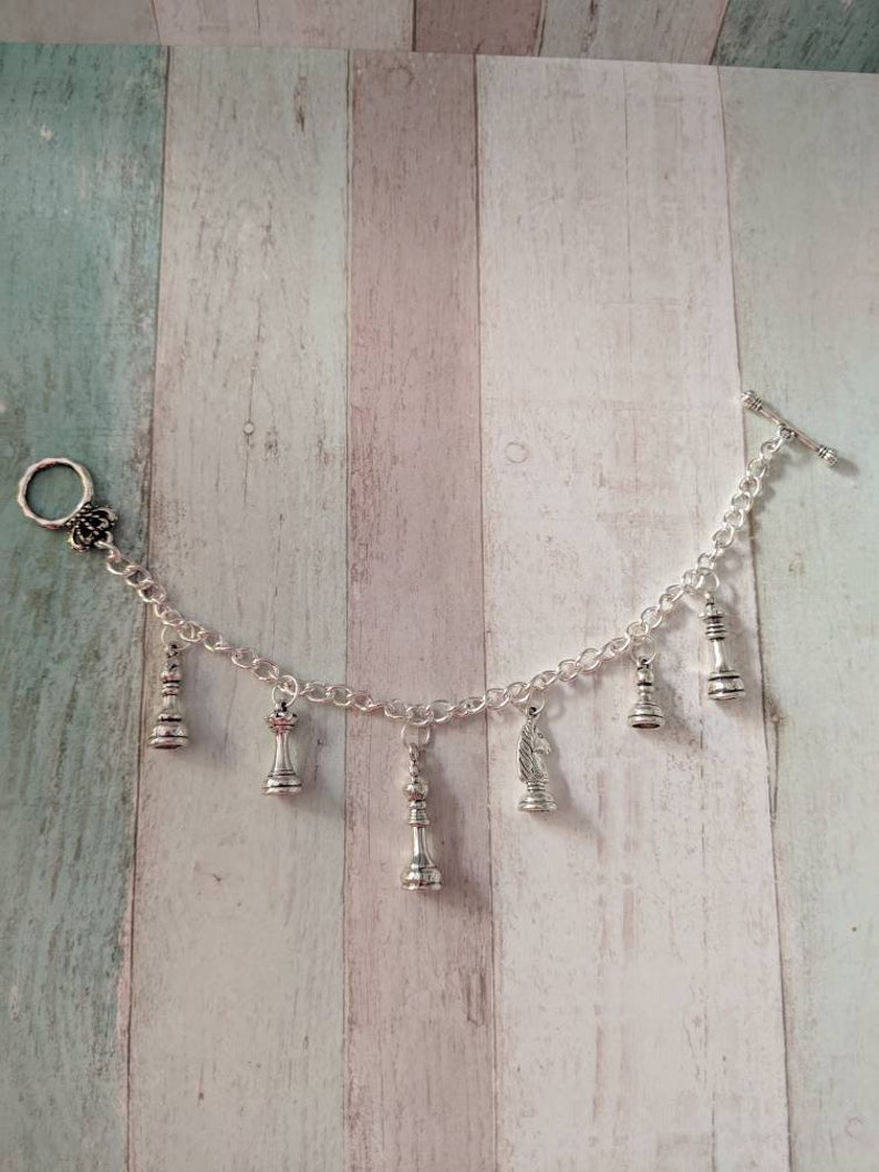 May include: A silver charm bracelet with a variety of chess piece charms, including a king, queen, knight, and pawn. The bracelet has a silver clasp and a silver toggle closure.