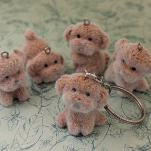 May include: A collection of small, plush dog keychains in various poses. The keychains are light brown with black eyes and noses, and each has a silver metal loop for attaching to a keyring. The background is a light green and white floral pattern.