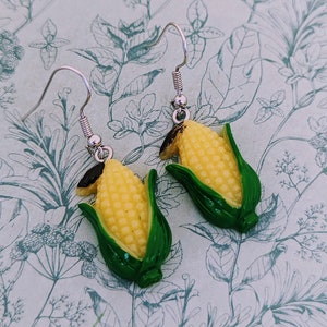 May include: A pair of silver earrings featuring a yellow corn on the cob with green husks. The corn is made of plastic and the earrings are dangle style.