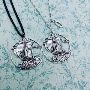 May include: Two silver necklaces with a ship pendant. The ship is detailed with a sail and waves. One necklace has a black cord and the other has a silver chain.