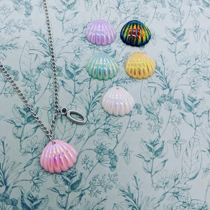 Puede incluir: Un collar de cadena plateada con un colgante de concha de mar rosa iridiscente. El collar está sobre un fondo azul claro con un patrón floral. Hay seis conchas iridiscentes adicionales en varios colores, incluyendo rosa, azul, amarillo y verde.