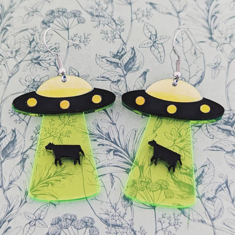 May include: A pair of green and black UFO earrings with a silhouette of a cow on each earring.