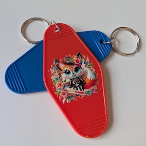 May include: Two motel-style keychains. One red keychain features a cartoon fox with a floral design and skeleton details. The other keychain is blue. Both have silver key rings.
