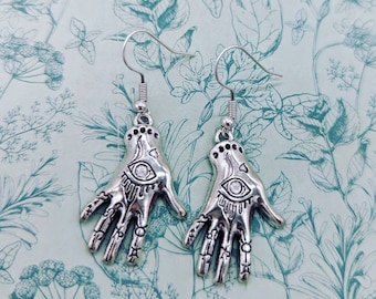 May include: Silver earrings with a hand design featuring an eye in the palm. The earrings are on a white background with a floral pattern.