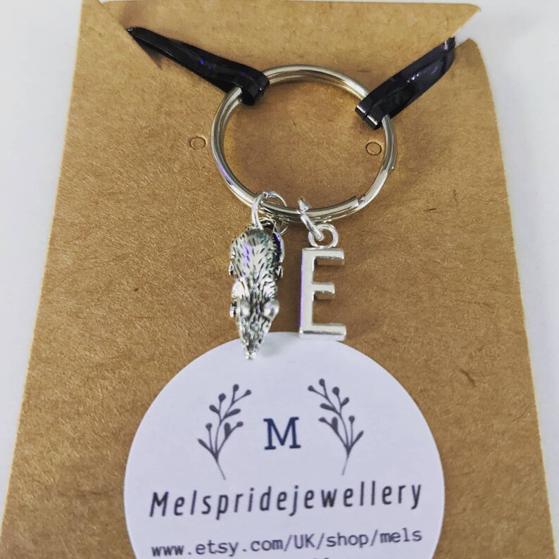 K&ouml;nnte beinhalten: Ein silberner Schl&uuml;sselanh&auml;nger mit einem kleinen Igel-Anh&auml;nger und dem Buchstaben E. Der Schl&uuml;sselanh&auml;nger ist an einer braunen Karte mit dem Text "Mels pridejewellery" und "www.etsy.com/UK/shop/mels" befestigt.