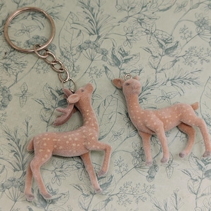 May include: Two deer figurines, one attached to a keychain, are displayed. The deer are light brown with white spots and a fuzzy texture. The keychain is silver. The background is a light green and white floral pattern.