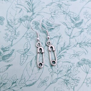 May include: A pair of silver safety pin earrings. The earrings are on a white background with a floral pattern.