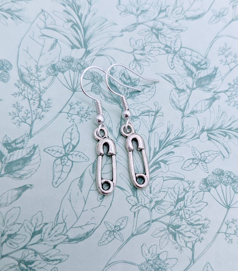 May include: A pair of silver safety pin earrings. The earrings are on a white background with a floral pattern.