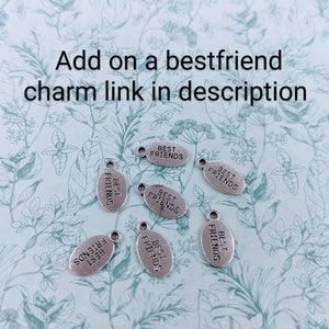 May include: Silver-colored oval charms with the words "BEST FRIENDS" are scattered across a light blue floral patterned surface. The charms have a small loop at the top for attaching to a bracelet or necklace. Text in the image reads "Add on a bestfriend charm link in description."
