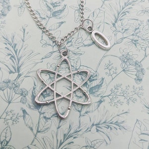 以下が含まれることがあります： 銀色のネックレス。原子の形をしたペンダントと、小さな楕円形のチャームが付いています。原子ペンダントには、周回する球があります。チェーンはカーブスタイルです。背景には、花柄の模様があります。