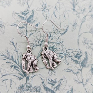 Puede incluir: Un par de pendientes colgantes de plata con un pequeño encanto de mono detallado en cada pendiente.