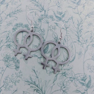 May include: A pair of silver earrings with a double female symbol design. The earrings are hanging from silver hooks.