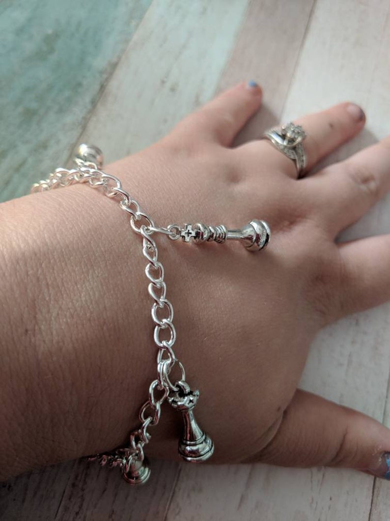 May include: A silver chain bracelet with three silver chess piece charms. The charms are a king, a queen, and a pawn.