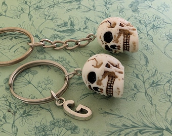 May include: Two keychains with skull charms. The skulls are off-white with brown accents, and the keychains have silver-colored rings and chains. One keychain has a silver letter "G" charm.