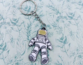 May include: A silver key chain with a white astronaut charm. The astronaut has a gold helmet and is wearing a spacesuit.