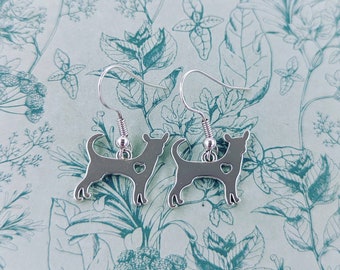 May include: Silver dangle earrings featuring a silhouette of a chihuahua with a heart cutout. The earrings are on a white background with a floral pattern.