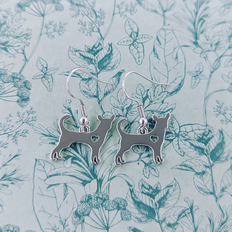 May include: Silver dangle earrings featuring a silhouette of a chihuahua with a heart cutout. The earrings are on a white background with a floral pattern.