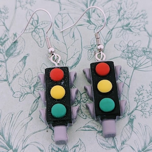 May include: A pair of novelty earrings shaped like traffic lights. Each earring features a black rectangular body with red, yellow, and green circular lights. The earrings have silver-colored hooks.