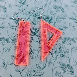 Può includere: Un paio di orecchini in acrilico rosa a forma di righello e triangolo. L'orecchino triangolare ha il testo "Back to School" stampato su di esso.