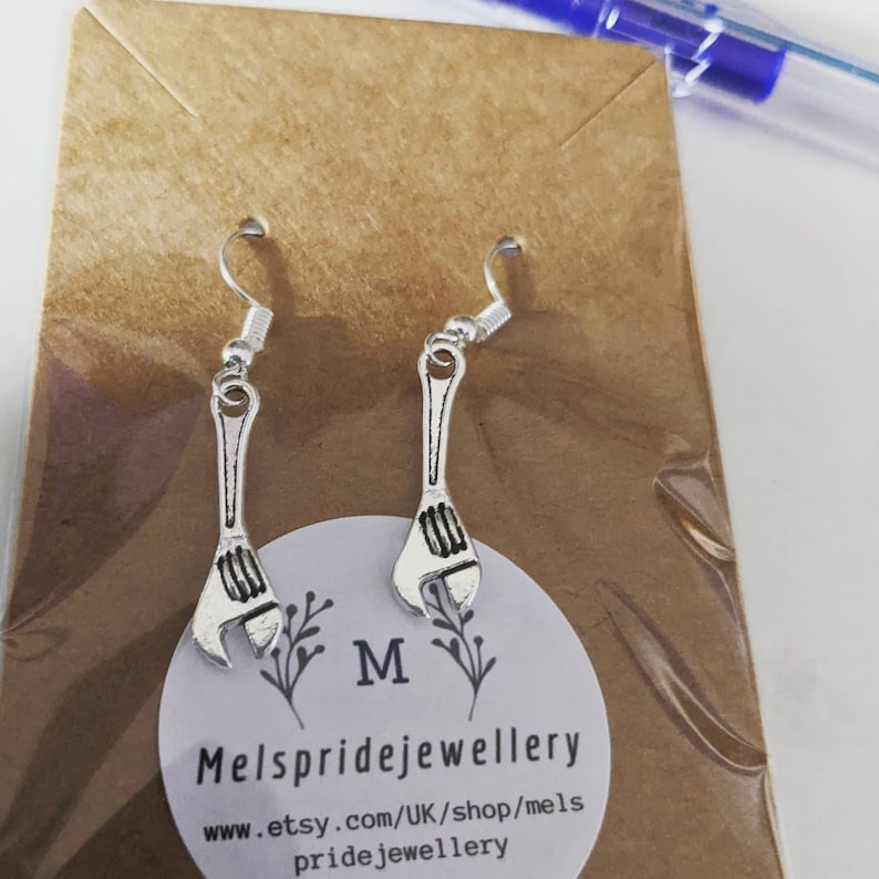 May include: Silver wrench earrings with a silver hook. The earrings are on a brown card with the text "Mels pridejewellery www.etsy.com/UK/shop/mels pridejewellery" printed on it.