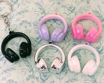Puede incluir: Una colección de colgantes de auriculares coloridos. Los colgantes son de varios colores, incluyendo negro, plateado, rosa, morado y blanco. Un colgante de auriculares negro está unido a un llavero. Los colgantes están sobre un fondo con estampado floral.