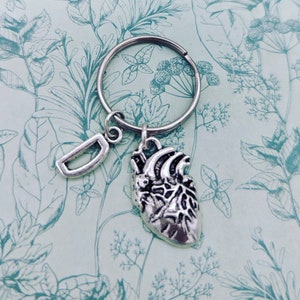 Op de afbeelding: Een zilverkleurige sleutelhanger met een bedel in de vorm van een menselijk hart en een kleine letter "D" bedel. De sleutelhanger ligt op een lichtblauwe achtergrond met een bloemenpatroon.