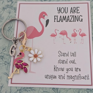 May include: A keyring featuring a pink flamingo charm, a white daisy charm, and a silver letter "G" charm. The keyring is placed next to a card with a flamingo illustration and the text "YOU ARE FLAMAZING".