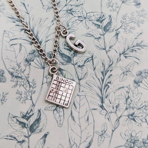Puede incluir: Un collar de plata con un dije de plata que dice "Bingo" y un dije de plata en forma de la letra "G".