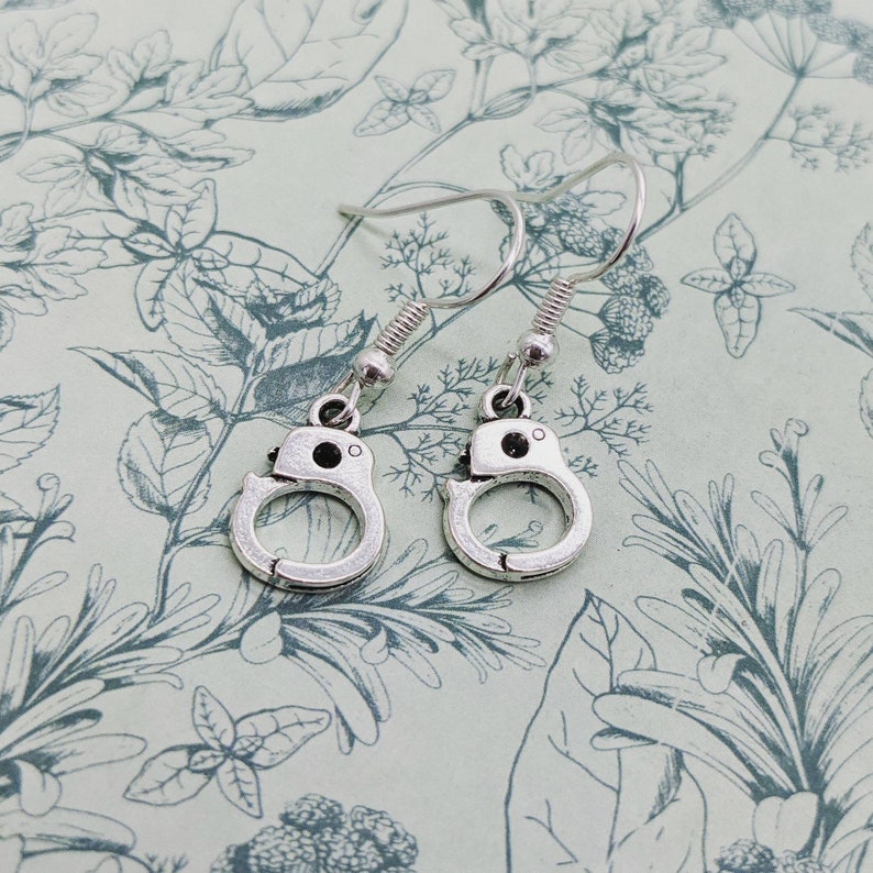 May include: A pair of silver dangle earrings featuring a handcuff design. The earrings have a simple, minimalist style.
