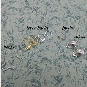 May include: A close-up of four different types of earring findings: lever backs, posts, clip-on, and hooks. The findings are silver and gold in color and are arranged on a floral patterned surface.
