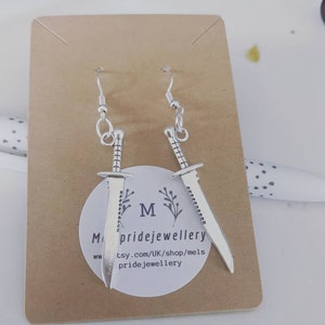 May include: Silver dangle earrings featuring a sword design. The earrings are displayed on a brown card with the text "Melspridejewellery" and a website address.