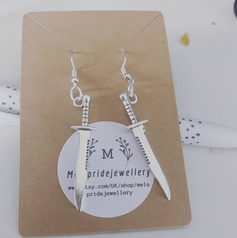 May include: Silver dangle earrings featuring a sword design. The earrings are displayed on a brown card with the text "Melspridejewellery" and a website address.