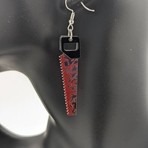 May include: A pair of silver earrings shaped like a saw with a black handle. The saw blade is red with a black and white pattern.