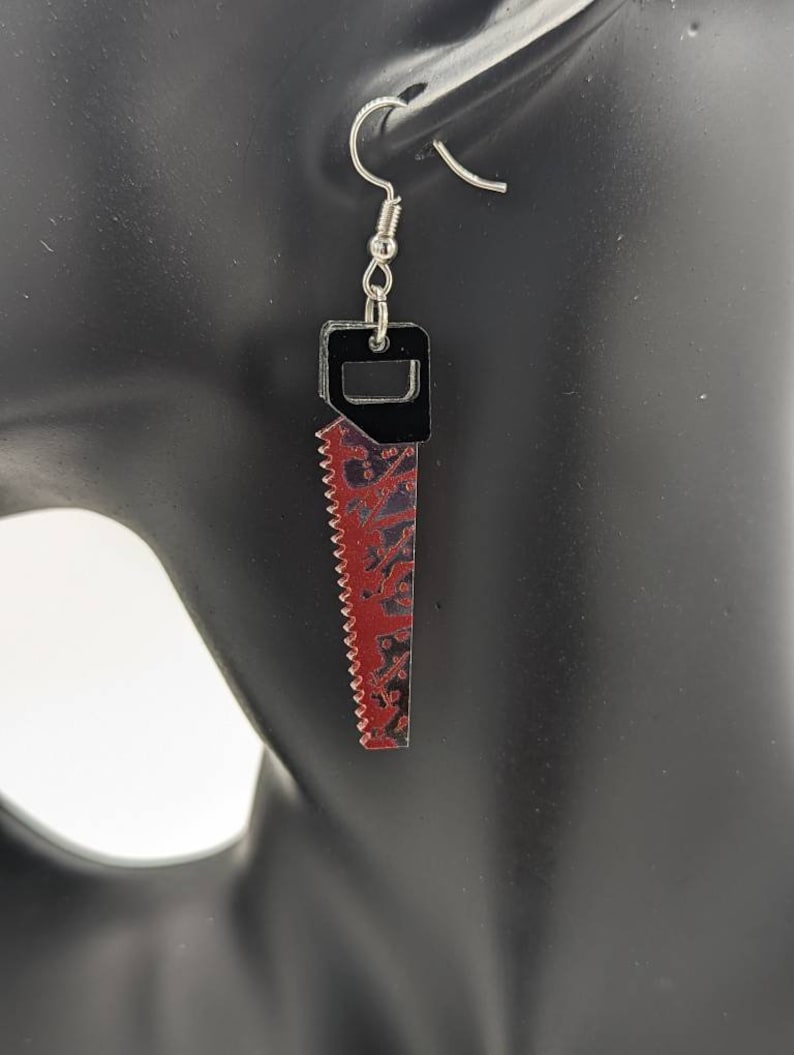 May include: A pair of silver earrings shaped like a saw with a black handle. The saw blade is red with a black and white pattern.