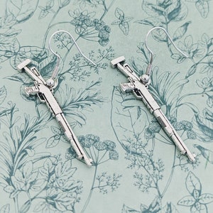 May include: A pair of silver shotgun earrings. The earrings are shaped like shotguns with detailed carvings. The earrings are hanging from silver hooks.