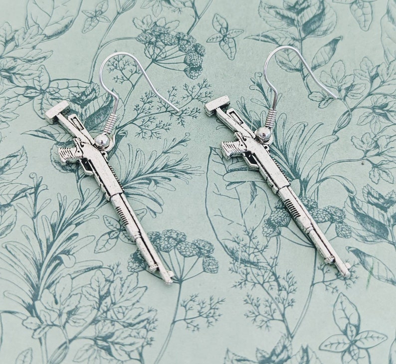 May include: A pair of silver shotgun earrings. The earrings are shaped like shotguns with detailed carvings. The earrings are hanging from silver hooks.