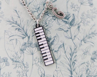 May include: A silver chain necklace with a piano key charm and a letter 'G' charm. The necklace is on a light blue background with a floral pattern.