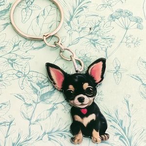 May include: A keychain featuring a black and tan Chihuahua dog with large ears and a red heart-shaped collar charm. The keychain has a silver-colored ring and chain. The background is a light green color with a floral pattern.