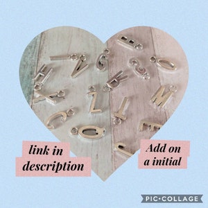 May include: Silver letter charms in the shape of the alphabet. The charms are arranged in a heart shape on a wooden surface. The text "link in description" and "Add on a initial" is visible in the image.