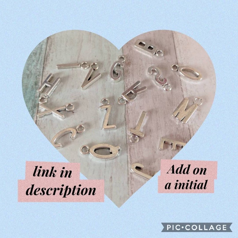 May include: Silver letter charms in the shape of the alphabet. The charms are arranged in a heart shape on a wooden surface. The text "link in description" and "Add on a initial" is visible in the image.