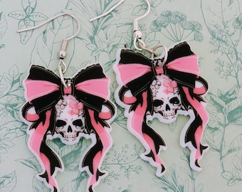 May include: A pair of pink and black skull earrings with a bow. The skull is decorated with pink flowers.