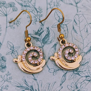 Può includere: Un paio di orecchini dorati con un design a spirale e strass trasparenti. Gli orecchini sono appesi su uno sfondo con un motivo floreale.
