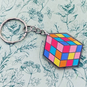 Puede incluir: Un llavero de cubo de Rubik colorido con un anillo y una cadena de metal plateado. El cubo presenta un patrón clásico de cuadrados rosas, azules, amarillos, naranjas y rojos brillantes. El llavero está sobre un fondo verde claro con un estampado floral.