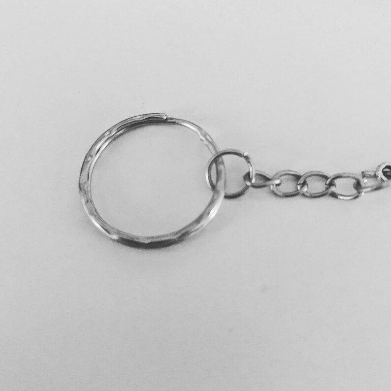 May include: Close-up of a silver-colored keychain with a textured, hammered finish on the large ring. A small ring connects to a chain of small, linked circles. The background is a plain, light surface.