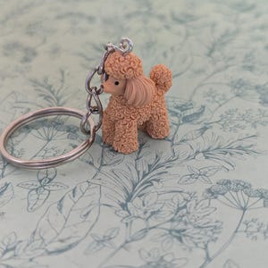 May include: A keychain featuring a small, light brown poodle figurine with a textured, curly coat. The poodle is attached to a silver-toned metal split ring by a short chain. The figurine has a smooth face, dark eyes, and a small nose.