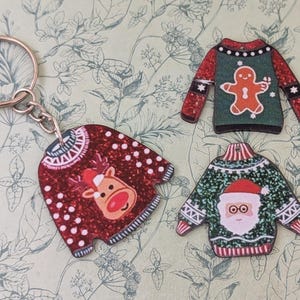 May include: A selection of Christmas jumper decorations. One is a keyring with a red reindeer design. Two other jumper ornaments feature a gingerbread man and Santa Claus. The jumpers have red, green, and white detailing.